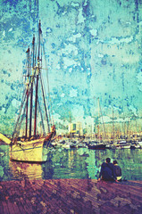 Young romantic couple sitting on wooden wharf at dusk in front of tall yachts at the port of Barcelona, Spain. Vintage grunge image textured with peeling paint.