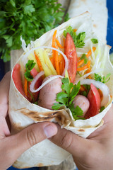Hot dog in hand on weight - sandwich with sausage in pita, Korean carrots, tomatoes, onions, parsley and pepper. Wooden background. Closeup