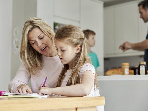 a woman and her daughter seated at a table in the kitchen looking at her homework. 