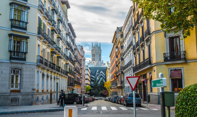 People cross the Street, in Madrid, Spain.  Pedestrian crossing, yield sign, colourful apartment buildings, mid-day street scene.  Urban life, parked cars in street. 
