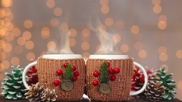 Closeup of two white steaming cups in handmade brown cute knitted covers with festive christmas decorations at blurry lights of orange garland background. Real time full hd video footage.