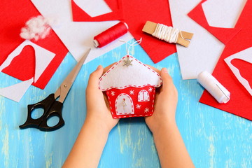Child sewed a felt Christmas tree house decoration. Child holds Christmas house decoration in his hands. Sewing tools and materials on blue wooden background. Easy crafts pfor children