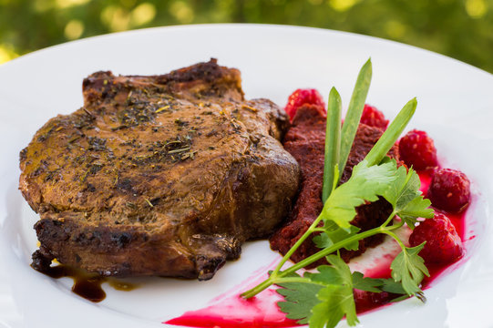 Fried Grilled pork steak on the bone with vegetables, mashed beetroot and berry sauce. Against the background of foliage. Close-up