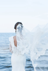 Naklejka premium The bride in a beautiful dress and vintage faie looking at the sea. Dress with open back. veil flies. Wedding dress with lace. Wedding photo concept. Bride. Art wedding photo