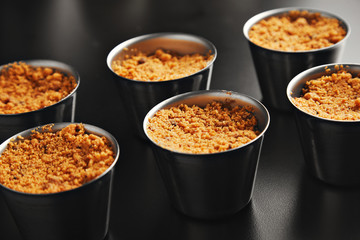 Close view on six portions of apple crumble dessert in individual steel cups on shiny black table