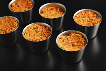 Set of six identical steel cups with hot brown crumble dessert on reflective black surface