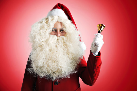 Santa Claus with huge pretty beard in red hat and coat ringing a golden bell on red background