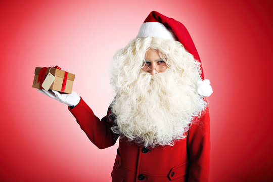 Trendy Santa in nice red clothes with huge white beard holding a present in craft cardboard box with red ribbon on red background