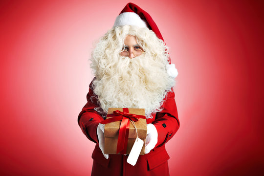Serious Santa Claus in a woolen coat, red hat and white mittens offering a present in a brown cardboard box with a red bow on red background