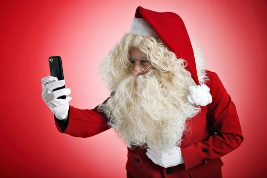 Santa is trying to see something on small screen of his new smartphone while taking photo or video, isolated on red