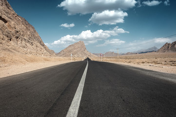 Iran - Empty tarred road in the desert with rocky mountains and blue cloudy sky