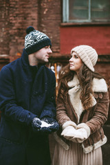 Young happy couple laughing and enjoying christmas street decorations with gifts in craft paper, cookies, cacao, handmade wooden table and red brick wall