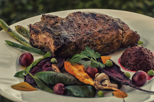 Fried Grilled pork steak on the bone with vegetables, mashed beetroot and berry sauce. Against the background of foliage. Close-up
