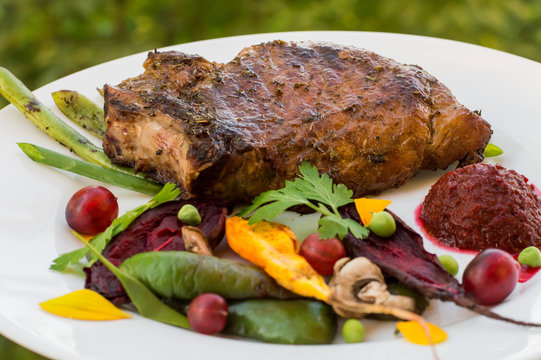 Fried Grilled pork steak on the bone with vegetables, mashed beetroot and berry sauce. Against the background of foliage. Close-up