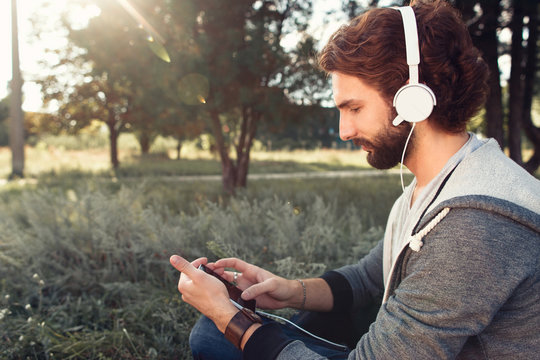 Profile of man in headphones using smartphone. Young guy choosing favorite music in mobile phone at nature, free space for text, sunlight flare. Relax, leisure, rest concept