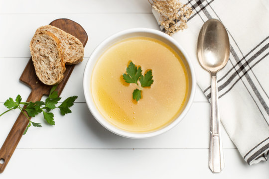 Sopa caldo dentro de un cuenco blanco y una cuchara de metal antigua junto a unas rodajas de pan y una servillleta sobre mesa de madera blanca. Vista superior