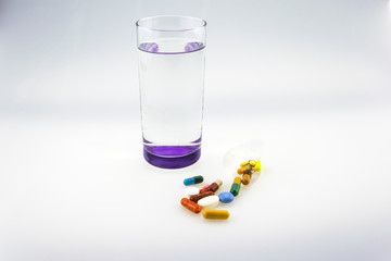 Many colored tablets and capsules near a glass of water with purple bottom isolated on white background.