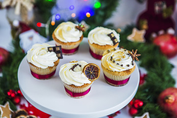 Пирожные, капкейки с сушеным лимоном и шоколадом на белой подставке на фоне новогодней зеленой гирлянды и новогодних огней.