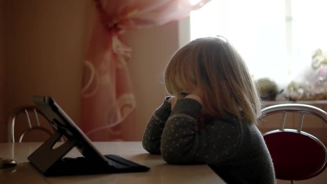 Pretty Little Girl Watches Cartoon Ontablet Computer Sits on the Kitchen Table