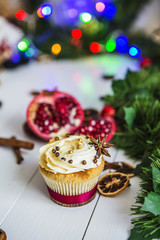 Пирожное с кремом, капкейк, мбирный пряник в виде звезды, разрезанный красный гранат, корица, сушеные лимоны лежат на белом деревянном столе на фоне зеленой новогодней гирлянды и новогодних огней.