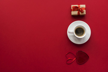  little white cup of aromatic black espresso, glass red heart and little gift wrapped in kraft paper on red vinous background 