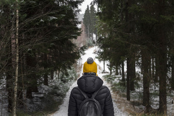 Fototapeta na wymiar alone young woman in black down jacket and yellow knitted hat with rucksack on the snow road in black forest during cold winter day in finland