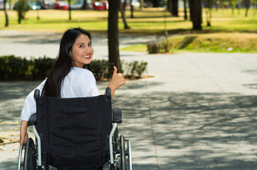 Young brunette woman sitting in wheelchair smiling with positive attitude, outdoors environment, physical recovery concept