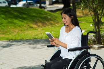 Young brunette woman sitting in wheelchair smiling with positive attitude, using mobile phone, outdoors environment, physical recovery concept