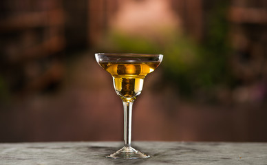 Classy cocktail glass sitting on grey surface, yellow liquid inside with blurry urban background
