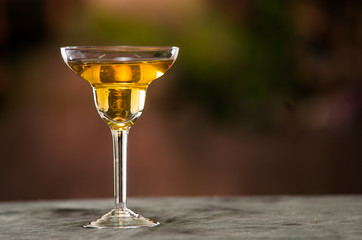 Classy cocktail glass sitting on grey surface, yellow liquid inside with blurry urban background