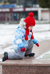 Little girl in a red cap gets on a marble slab in winter