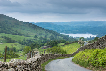 Fototapeta na wymiar Lake District, England - May 30, 2012: Rural road with stone walls on the side meanders through the landscape. Shot from above shows lake downhill. Green meadows, forests and ferns