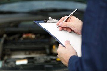 Mechanic standing in front of an open car hood with clipboard and pen. Closeup