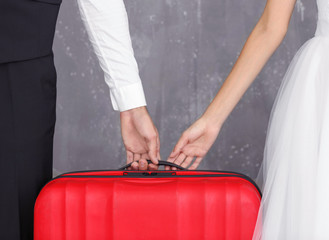 Groom and bride holding red suitcase on grey background