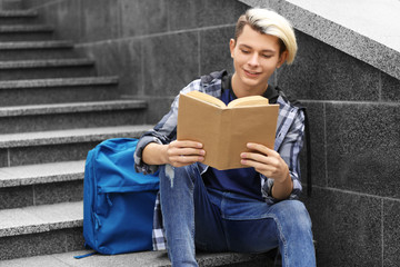 Cute teenage boy with book sitting on stairs outdoors