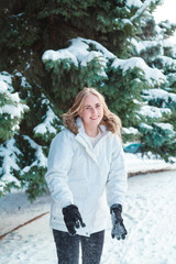 On a cold winter day young beautiful cheerful woman with long blond hair in a white jacket plays with snow against the backdrop of snow-covered trees and cheerfully laughs