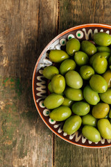 Green olives in brown bowl on wooden background. Top view. Copy space.
