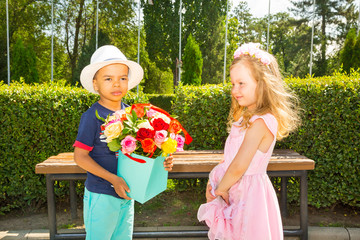 Black african american boy kid gives flowers to girl child on birthday. Little adorable children in park. Childhood and love.