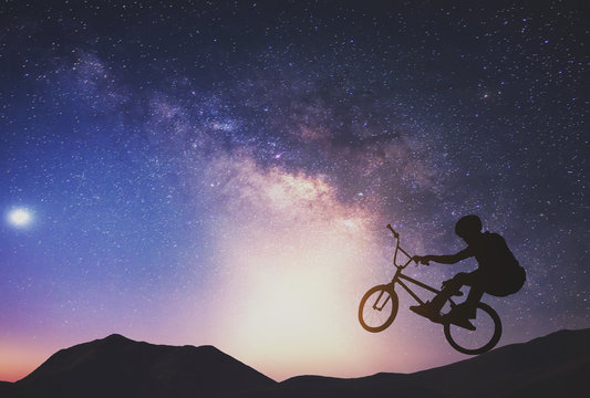 Man Riding A Bike Performing A Trick Against On Mountain With Milky Way.