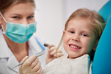 Happy smiling child sitting on dental chair ready to cure teeth. Portrait of woman dentist in mask with tools treating patient. 