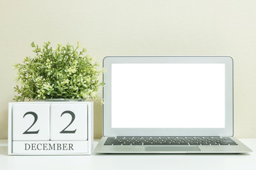 White wooden calendar with black 22 december word with white blank space at center of computer notebook on white wood desk and cream wallpaper textured background , selective focus at the calendar