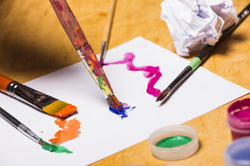 Hands of the artist  in bright colored paint with brush on wooden table. Woman draws on a sheet of paper