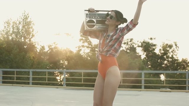 Smiling joyful woman in sunglasses and red swimsuit holding old record player and dancing on the open road outdoors