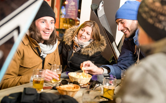 Happy friends drinking beer and eating chips - Cheerful people having fun at bar restaurant by skiing resort with snow equipment - Friendship concept on warm night filter with focus on young woman