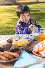 An Asian cute young boy eating breakfast outdoor at camping site in nature sunlight with a tent at the background.