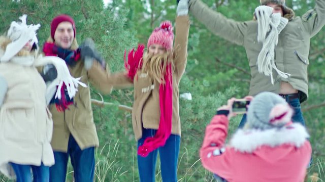 Young friends holding hands and jumping on snow in forest at winter day while girl taking picture of them with smart phone