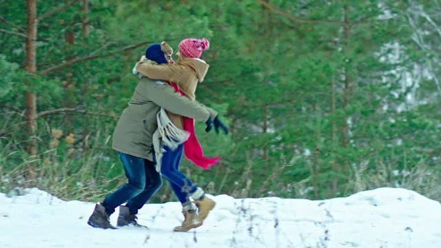 Young happy couple running through snowy forest at cold winter day. Boyfriend taking girl on his arms and spinning around in slow motion