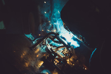 Industrial worker welding round pipe on a work table, producing blue smoke, yellow sparks and reflections