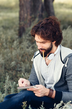 Close-up of thoughtful man with notebook biting pencil. Attractive bearded guy writing, drawing, counting in his sketchbook at nature, copy space.