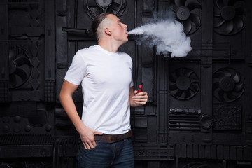 A man in a white T-shirt and jeans on a black background vaping and releases a cloud of steam.